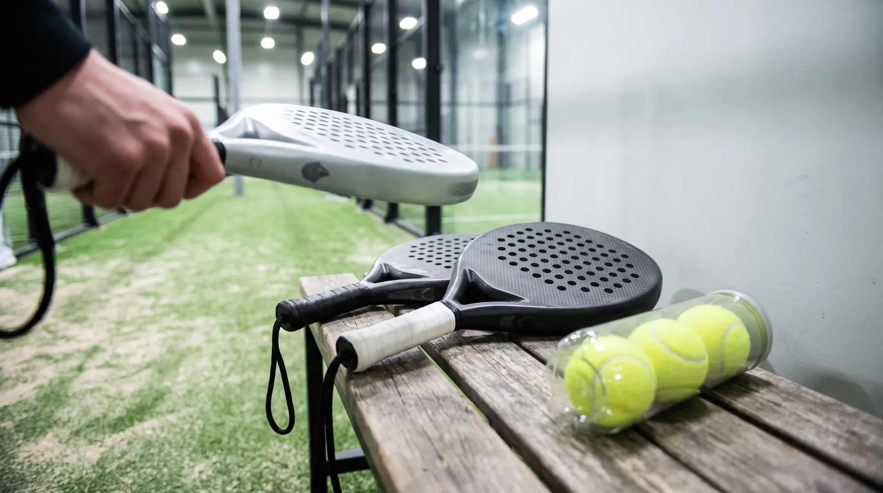 Padel rackets and a tube of balls on a courtside bench, a hand holding a lighter racket